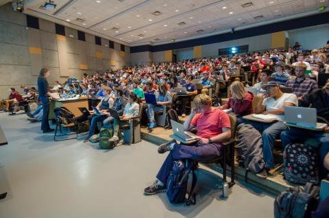 Students are seated in a large lecture classroom taking notes on laptop computers.
